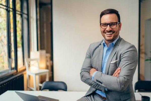 Portrait of young happy businessman wearing grey suit and blue shirt standing in his office and smiling with arms crossed
